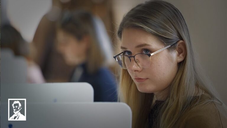 Woman with glasses focused on a laptop at a desk while others work in background
