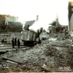 Workers stand near an industrial building demolition site with debris and smoke.