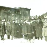 Men in overcoats and hats stand in a line outside buildings.