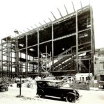 Large steel-framed building under construction or demolition with several vintage cars in front.