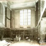 Interior of an old theater with tall windows, scaffolding, and stacked boards.