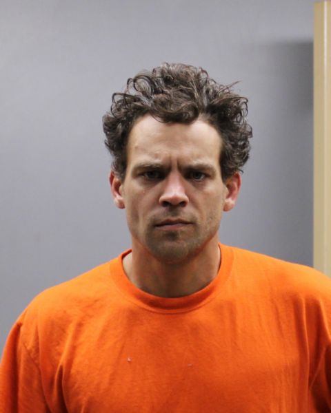 A man with curly hair wearing an orange prison-style shirt against a plain wall