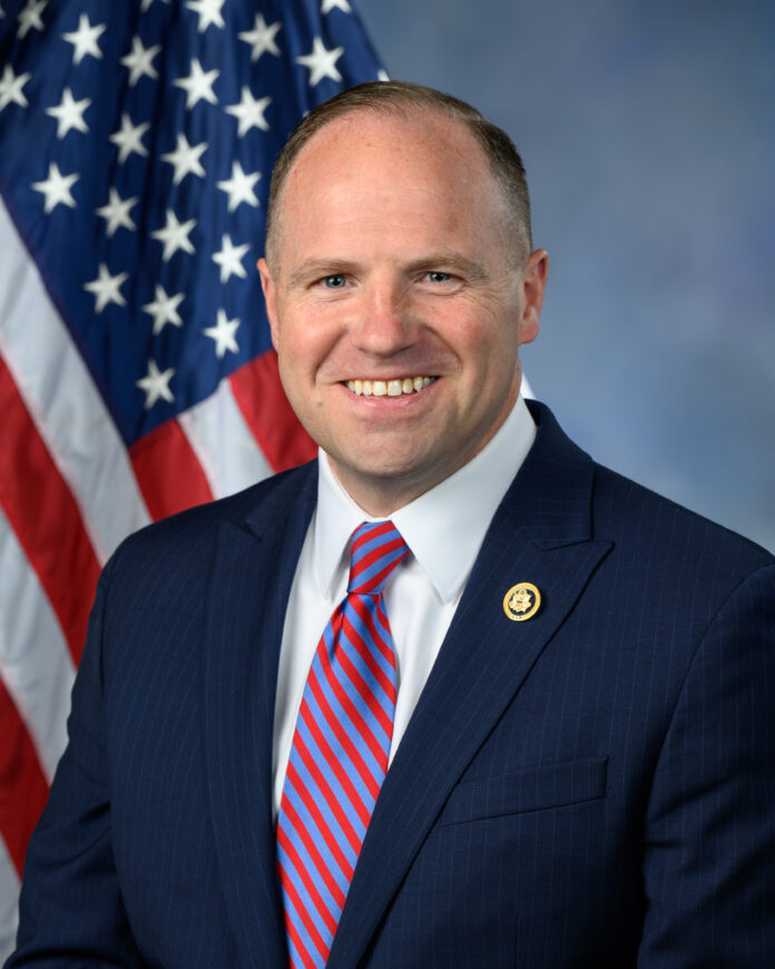 Smiling man in dark suit with striped tie, standing in front of U.S. flag