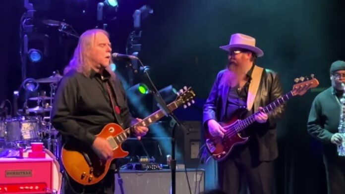 Band members sing and play electric guitars under purple stage lights.