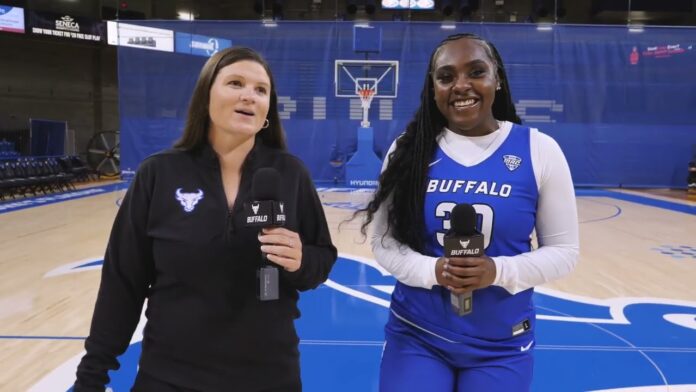 Two women stand courtside holding microphones in front of a blue basketball hoop backdrop.