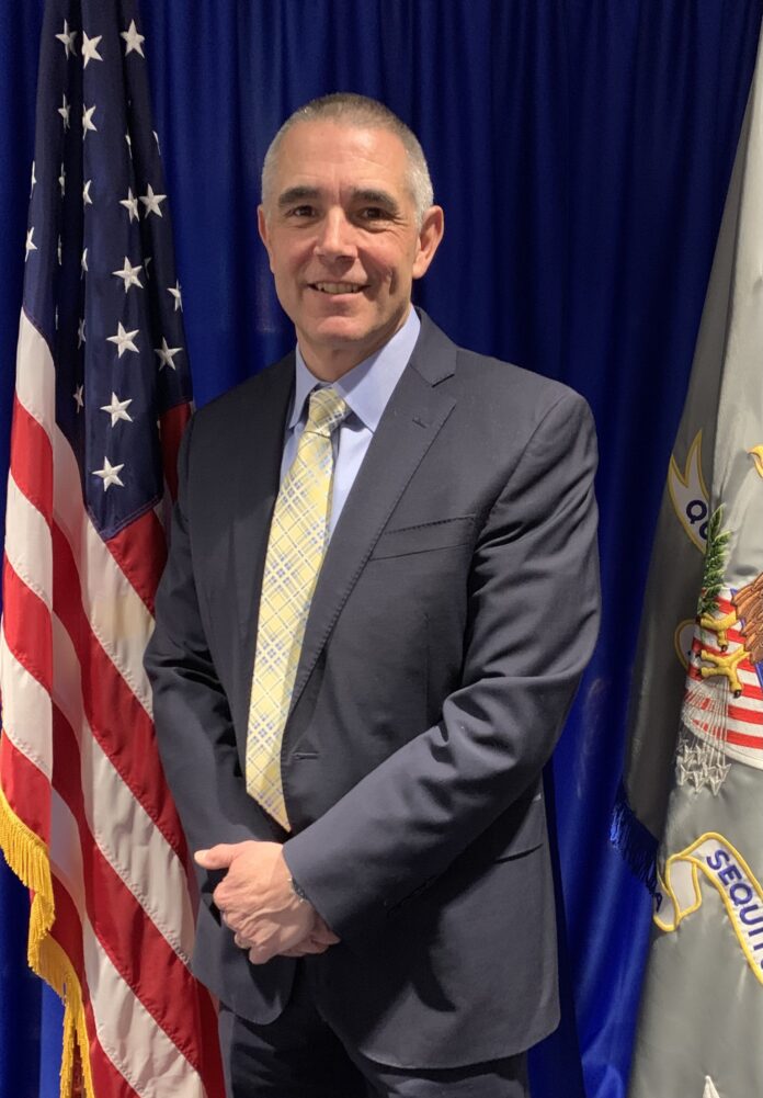 Man in a suit standing in front of U.S. flags and a blue backdrop, smiling.