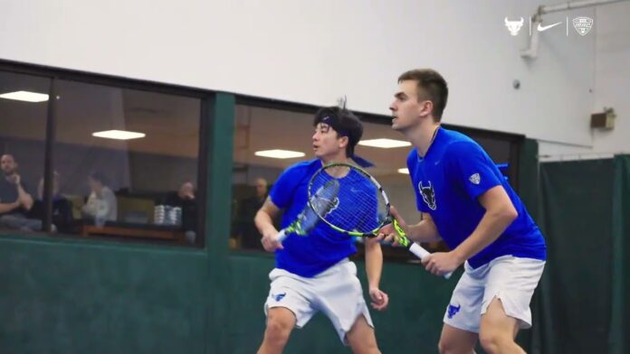 Two athletes in blue tennis shirts play a match on an indoor court.