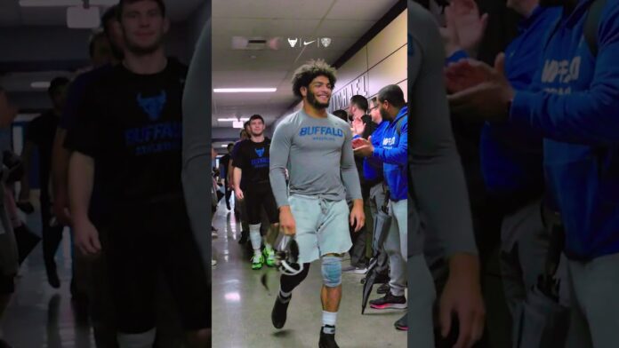 Athlete in gray Buffalo gear walks down a hallway as others in blue cheer nearby.