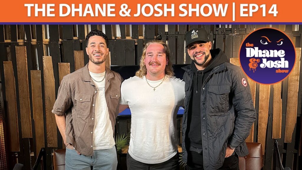 Three hosts stand outdoors in front of wooden boards; show logo reads “The Dhane & Josh Show | EP14”.