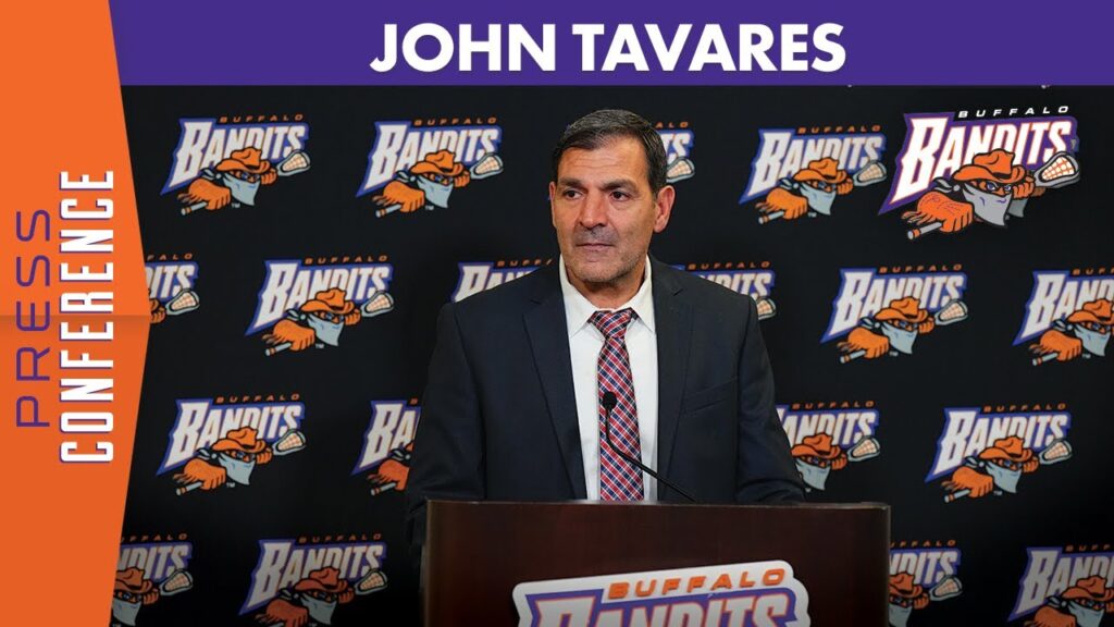 Man in suit at a podium with a Buffalo Bandits logo backdrop behind him.
