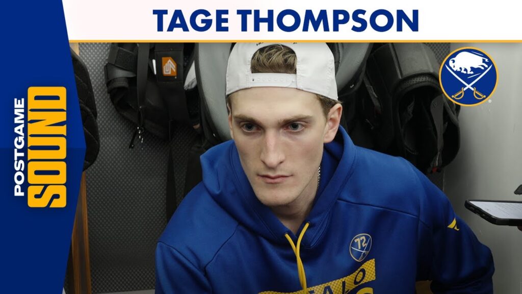 Tage Thompson seated in a hockey locker room wearing a blue hoodie and white cap.