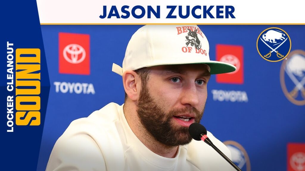Man in a white cap speaks into a microphone at a press backdrop