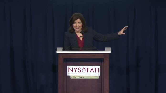 Woman speaking at a podium with two microphones, raising one hand before a NYSAFAH sign