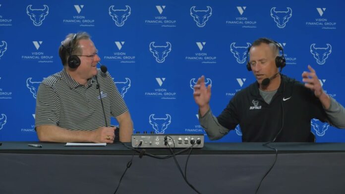 Two men wearing headsets speak into microphones at a podcast table with a blue sponsor backdrop.