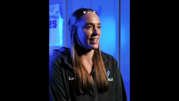 Woman in dark jacket seated in a blue-lit studio interview setup, speaking toward camera