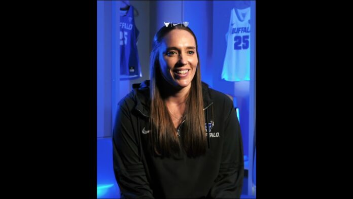 Woman in a blue-lit studio wearing a basketball-style cap and dark jacket, smiling at camera.