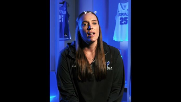 A woman in a black athletic jacket sits in front of blue-lit sports banners.