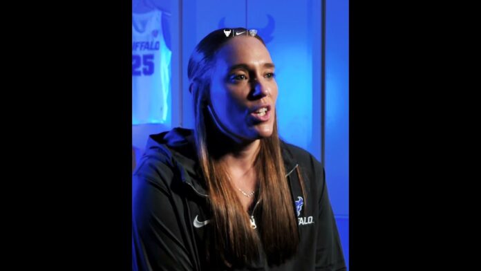 Woman in a dark sports jacket speaking in front of a blue backdrop with logos
