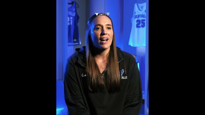 A woman in a dark athletic top speaks in a studio with blue lighting and sports banners behind her.