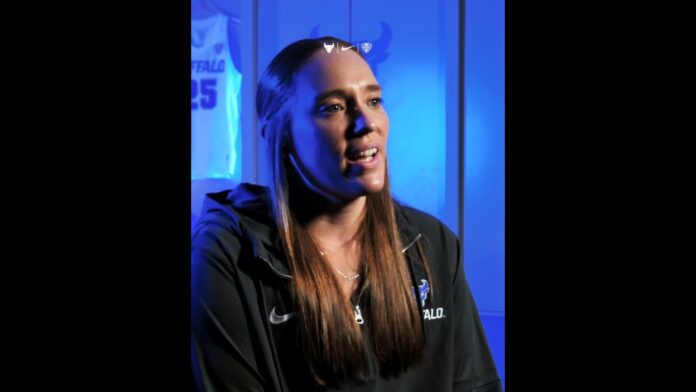 Woman in a dark zip-up jacket speaks during an interview in blue studio lighting