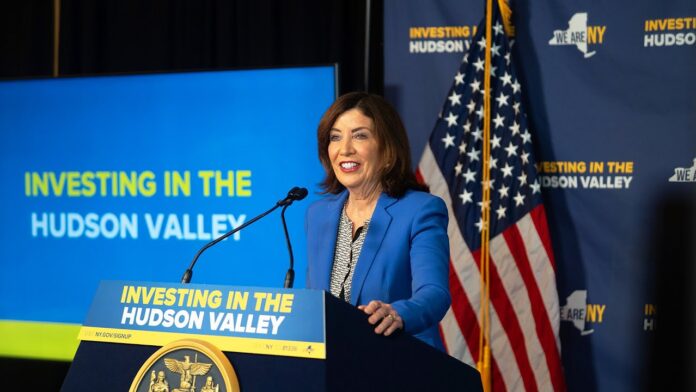 Person in blue blazer speaks at a podium with flags and a “Investing in the Hudson Valley” backdrop.