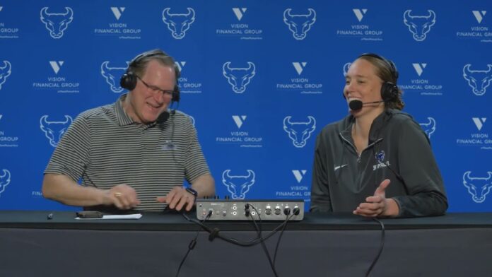 Two people wearing headsets with microphones sit at a table in front of a blue backdrop.