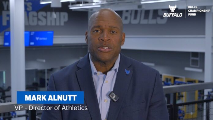 Man in a suit seated indoors with “Bulls Championship Fund” signage in the background.